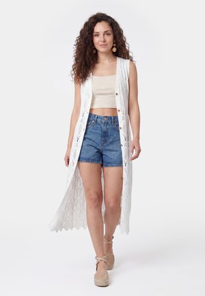 Femme aux cheveux bouclés portant un crop top beige, un long gilet blanc sans manches en maille, un short en denim et des espadrilles beiges, marchant vers l'avant.
