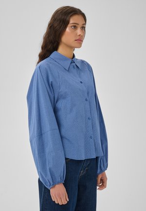 Femme debout en chemise bleue boutonnée et jean foncé, tournée vers la gauche devant un fond uni clair, avec une expression faciale neutre.