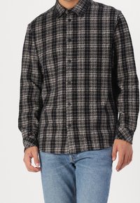 Homme portant une chemise à carreaux noire et blanche à boutons avec manches longues et un jean bleu clair, debout les mains détendues le long du corps.