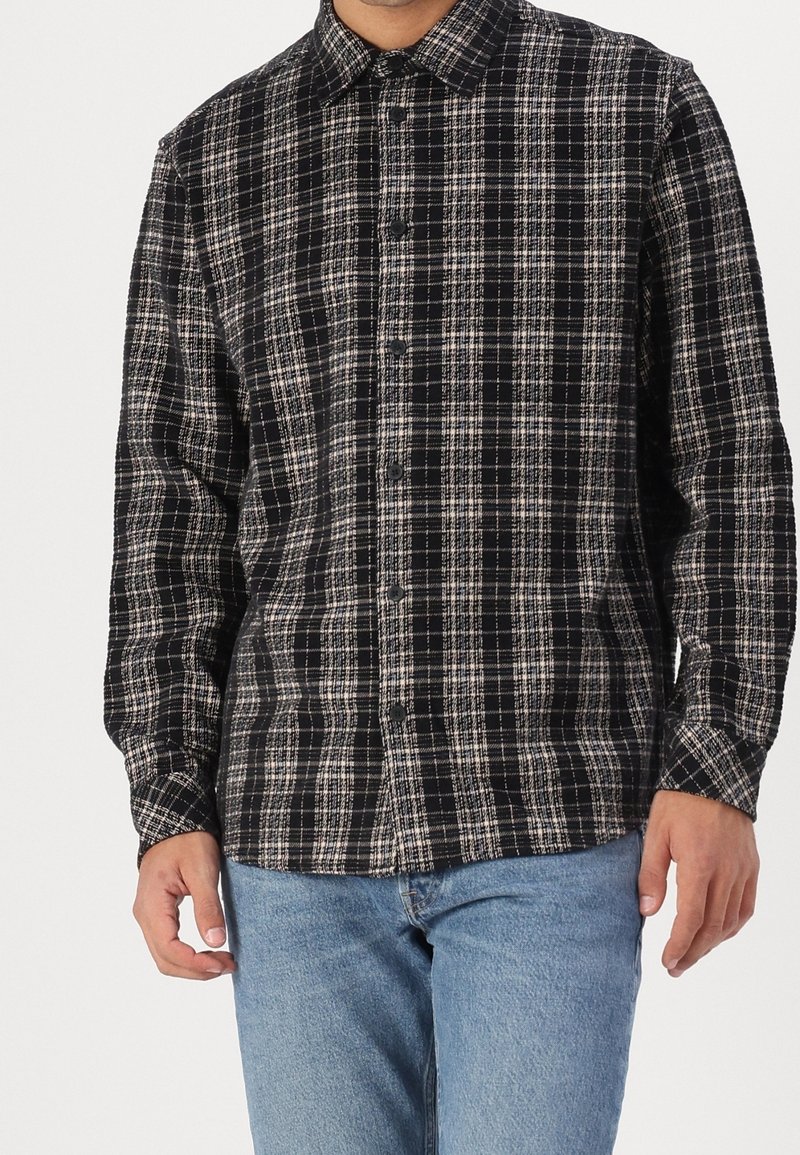 Homme portant une chemise à carreaux noire et blanche à boutons avec manches longues et un jean bleu clair, debout les mains détendues le long du corps.