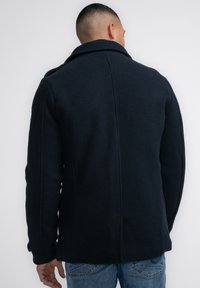 Abrigo de mezcla de lana en color azul marino con un amplio cuello y un acabado texturizado. Presenta costuras verticales y puños abotonados, combinado con jeans azules.
