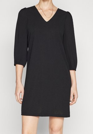 Kobieta ubrana w czarną sukienkę do kolan z plisowanymi rękawami 3/4 i dekoltem w kształcie litery V, ozdobioną cienkim złotym naszyjnikiem.