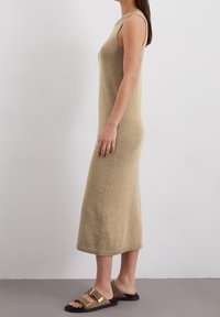 Abito lungo senza maniche color beige in maglina con silhouette aderente, scollo a coste e orlo. Abbinato a sandali marroni con una fibbia dorata.