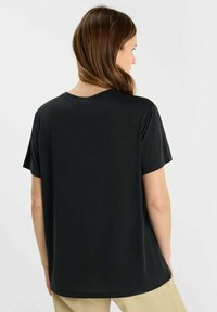 Vrouw met bruin haar, gekleed in een effen zwarte T-shirt met korte mouwen en beige broeken, met de rug naar de camera, tegen een neutrale achtergrond.