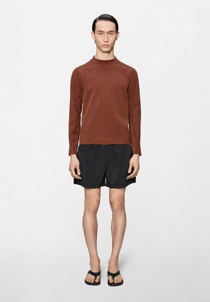 Αρσενικό μοντέλο στέκεται κοιτάζοντας μπροστά φορώντας καφέ μακρυμάνικη μπλούζα, μαύρο σορτς και μαύρα σανδάλια, πάνω σε απλό λευκό φόντο.