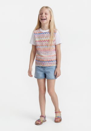 Fată zâmbitoare cu păr blond lung, purtând un top colorat fără mâneci cu model zigzag, tricou alb, pantaloni scurți din denim și sandale multicolore.