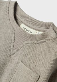 Maglione grigio chiaro con finitura texture, design a V, collo a costine e tasca frontale. L'etichetta riporta "Lil'Atelier."