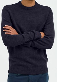 Person mit mittelbrauner Haut, der einen dunkelgrauen Strickpullover und dunkelblaue Hosen trägt, mit verschränkten Armen vor der Brust.