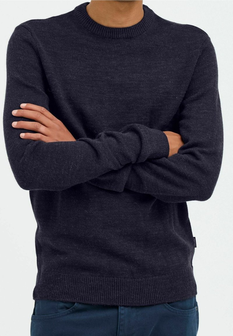 Person mit mittelbrauner Haut, der einen dunkelgrauen Strickpullover und dunkelblaue Hosen trägt, mit verschränkten Armen vor der Brust.