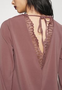 T-shirt de manga longa em malva com um decote em V nas costas, apresentando aplicação de renda e fechamento com laço. A textura suave com detalhes delicados realça a elegância.