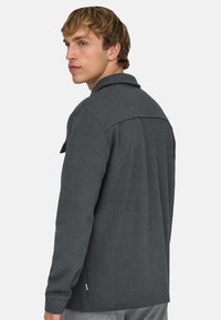 Junger Mann mit kurzem blondem Haar, der ein dunkelgraues Langarmshirt trägt, seitlich blickend mit neutralem Gesichtsausdruck vor einem einfarbigen Hintergrund.