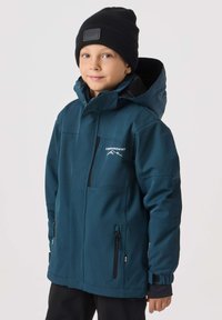 Morska miękka kurtka softshell z kapturem i wieloma kieszeniami na zamek. Ma czarne akcenty i logo na piersi. Noszona z czarną czapką beanie.