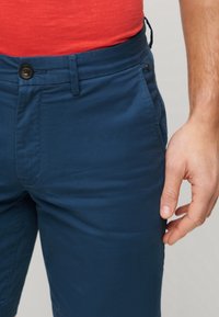 Pantalon en coton bleu avec fermeture à boutons, poches avant et texture lisse. Gros plan sur le tissu et une main saisissant la taille.