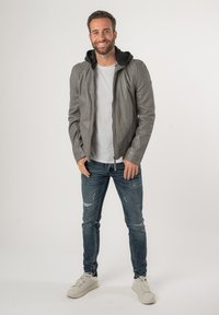 Grijze leren jas met zwarte capuchon, aansluitend ontwerp, ritssluiting, gecombineerd met versleten blauwe jeans en witte sneakers. Eenvoudige achtergrond.