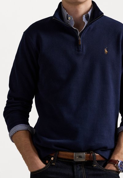 Man wearing navy quarter-zip sweater over blue and white striped button-up shirt, with hands in jeans pockets and brown leather belt.