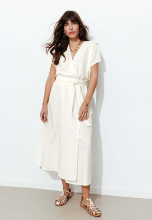 Femme aux longs cheveux bruns portant une robe portefeuille blanche à manches courtes, des bijoux en or et des sandales plates beiges à brides, debout contre un mur blanc.