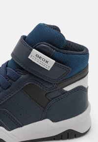 Modrá vysoká teniska s koženým a síťovaným svrškem, nastavitelné suché zipy, perforovaný boční detail a bílá gumová podešev. Obsahuje branding GEox.