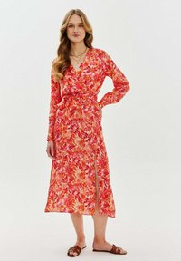 Dlhé kvetinové šaty v živých odtieňoch ružovej a oranžovej s hlbokým výstrihom do V, dlhými rukávmi a bočným rozparkom. Tkanina vyzerá ľahko a vzdušne.