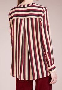 Chemise à manches longues avec rayures verticales crème, bordeau et noires. Détail de pli au dos pour plus de volume. Poignets boutonnés avec petits boutons blancs.
