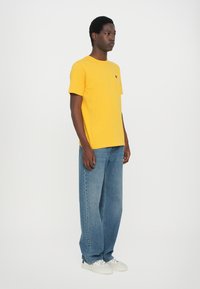 Tricou din bumbac galben, cu guler rotund, manșete scurte și un mic logo negru pe piept, asociat cu blugi drepti de culoare albastru deschis.
