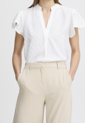 Femme portant un chemisier blanc à manches courtes texturé avec des manches volantées, rentrée dans un pantalon beige taille haute, les mains dans les poches.