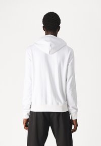 Vit bomullshuvtröja med en stor huva, ribbade ärmslut och rak nederkant. Baksidan är slät med synliga sömdetaljer.