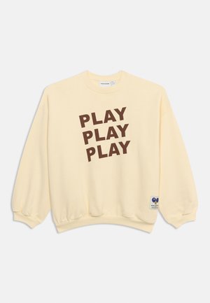 Kermanvärinen collegepaita, jossa on kolme ruskeaa ”PLAY”-sanaa pinottuna vinosti edessä ja pieni sininen sydämen muotoinen paikka helman lähellä.