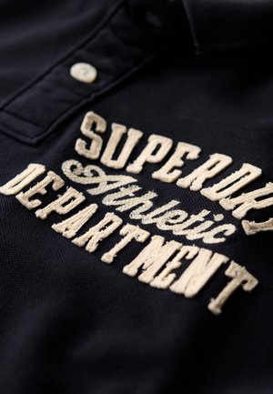 Polo-shirt marine avec une broderie texturée couleur crème indiquant "SUPERDRY Athletic DEPARTMENT", et une patte de boutonnage à trois boutons.