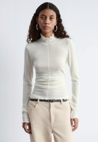 Langärmliges Oberteil mit hohem Kragen in Cremefarbe, taillierte Silhouette, Rüschensaum und cuffs. Kombiniert mit beigen gerippten Hosen und einem Gürtel.