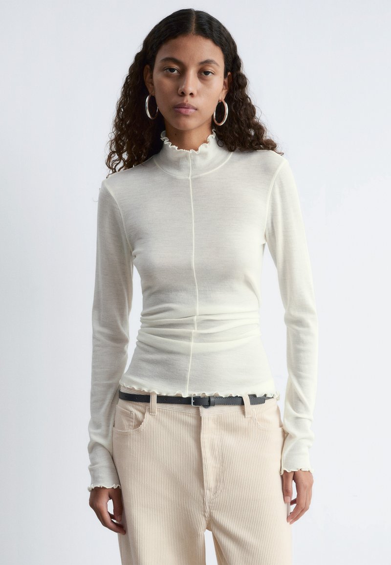 Langärmliges Oberteil mit hohem Kragen in Cremefarbe, taillierte Silhouette, Rüschensaum und cuffs. Kombiniert mit beigen gerippten Hosen und einem Gürtel.