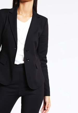 Frau trägt einen maßgeschneiderten schwarzen Blazer über einer weißen Bluse, kombiniert mit schwarzen, eng anliegenden Hosen, stehend vor einem schlichten weißen Hintergrund.