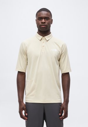 Jeune homme noir debout, portant un polo beige à manches courtes et un pantalon foncé, face à un fond blanc uni.