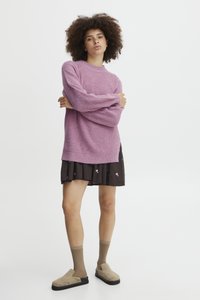 Różowy sweter ribbed z długimi rękawami, noszony na ciemnej spódnicy ozdobionej małymi wzorami kwiatowymi, połączony z beżowymi butami wsuwanymi i brązowymi skarpetkami.