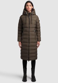 Lang, olivengrøn puffjakke med hætte, vertikal quilting, front lynlås og sidelommer. Iført over sorte leggings og sorte ankelstøvler.