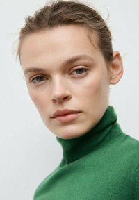 Pulover verde cu guler înalt, textură moale, croială mulată și accente cu dungi la guler, încadrând un chip cu tonuri neutre.