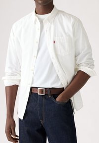 Camisa blanca de botón con mangas enrolladas, combinada con un cinturón de cuero marrón y jeans de mezclilla azul oscuro. Incluye un bolsillo en el pecho y hebilla plateada.