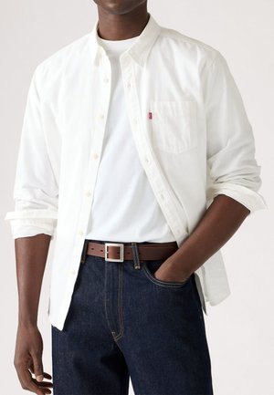 Camisa blanca de botón con mangas enrolladas, combinada con un cinturón de cuero marrón y jeans de mezclilla azul oscuro. Incluye un bolsillo en el pecho y hebilla plateada.