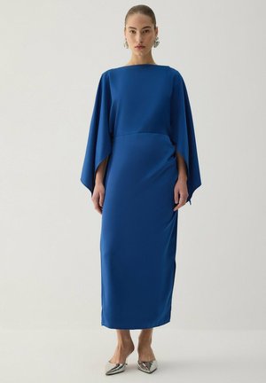 Femme debout dans une longue robe bleue à manches larges, aux talons aiguilles argentés et grandes boucles d’oreilles argentées, sur un fond uni.