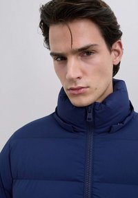 Joven con cabello oscuro que lleva una chaqueta acolchada azul marino con cremallera, mirando ligeramente de lado con una expresión neutral, fondo claro y sencillo.