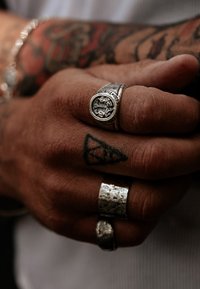 Sidabriniai žiedai ant rankos, viename žiede pavaizduota detali emblema, kiti - su tekstūruotais paviršiais, o ant odos matomi tatuiruotės.