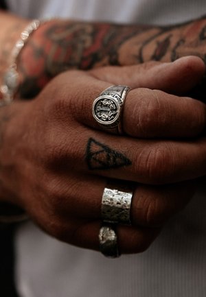 Silver rings on a hand, featuring a detailed emblem on one ring, textured surfaces on others, and visible tattoos on the skin.