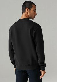 Sudadera negra con cuello redondo y puños acanalados, que presenta una textura suave y un diseño liso, llevada sobre pantalones oscuros.