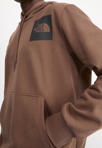 Hnědá mikina s kapucí a přední kapsou, s černým logem "The North Face" na levé hrudi, nošena člověkem s ohnutou paží v pase.