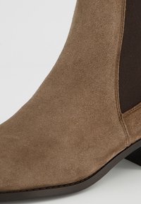 Hnědá semišová Chelsea bota s elastickým bočním panelem a černou podrážkou, zobrazená zblízka na světlém pozadí.