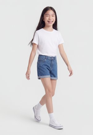 Smilende pige, der går, iført hvid t-shirt, opkørte denimshorts, hvide sokker og hvide sneakers mod en ensfarvet lys baggrund.