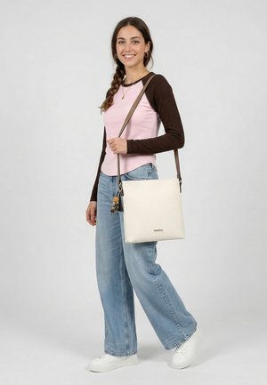 Noor naeratav naine, seljas roosa ja pruun pika varrukaga särk, laiad sinised teksapüksid, valged tossud ning kandmas valget õlakotti.