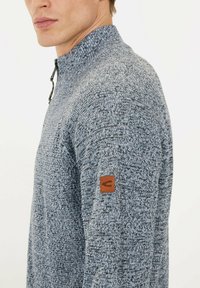 Gestrickter Pullover in Blau- und Grautönen mit hohem Kragen und Reißverschluss. Verfügt über ein strukturiertes Muster und ein braunes Leder-Logopatch am Ärmel.