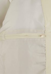 Light beige jacket featuring smooth fabric, a zippered pocket on the left side, and tonal stitching along the seams.