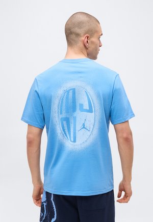 Muž nosí svetlomodré tričko s veľkou abstraktnou kruhovou grafikou a malým logom Jumpman na chrbte, otočený chrbtom k fotoaparátu.