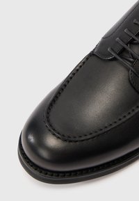 Nærbillede af en sort læder dressko med detaljeret syning og snørebånd på hvid baggrund.
